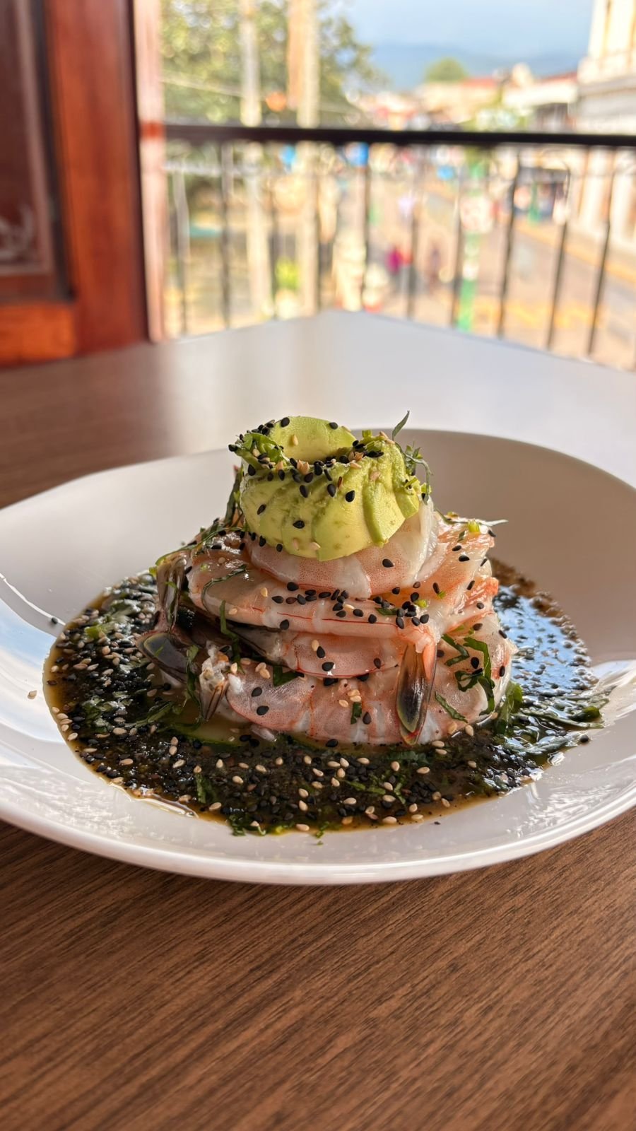 Gourmet plated dish with stacked smoked salmon, avocado, and seaweed on white plate with city view balcony backdrop